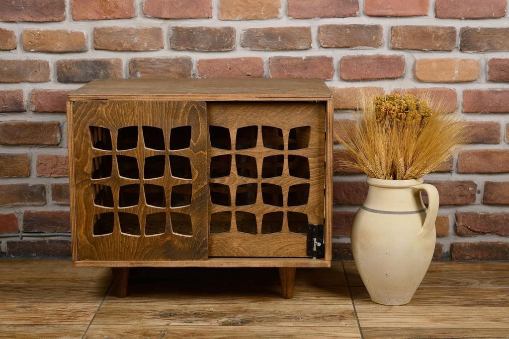 Wooden dog crate with lattice design next to a vase with dried plants against a brick wall.