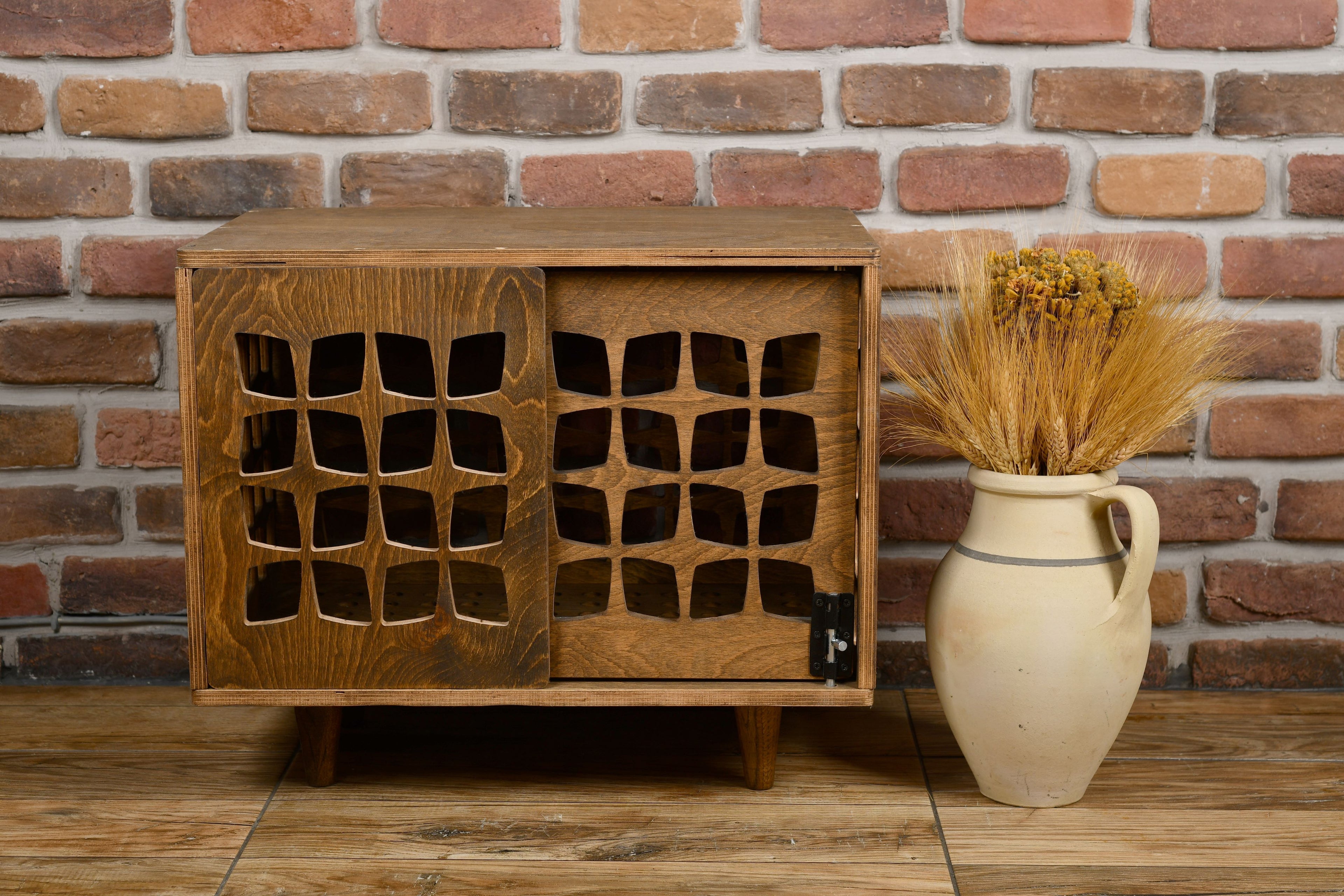 Wooden dog crate with lattice design next to a vase with dried plants against a brick wall.