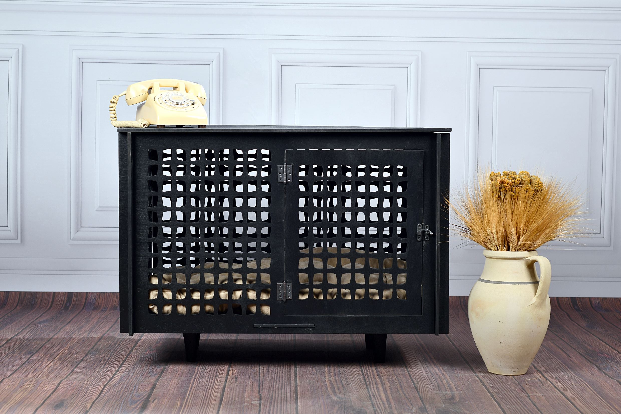 Black wooden dog crate with a vase of dried plants on a wooden floor against a light blue wall.
