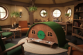 A hobbit-style pet house with a curved green grass roof, wooden door frame, and elevated wooden platform with two metal bowls, placed in a cozy fantasy-themed room with round windows, chairs, and shelves.